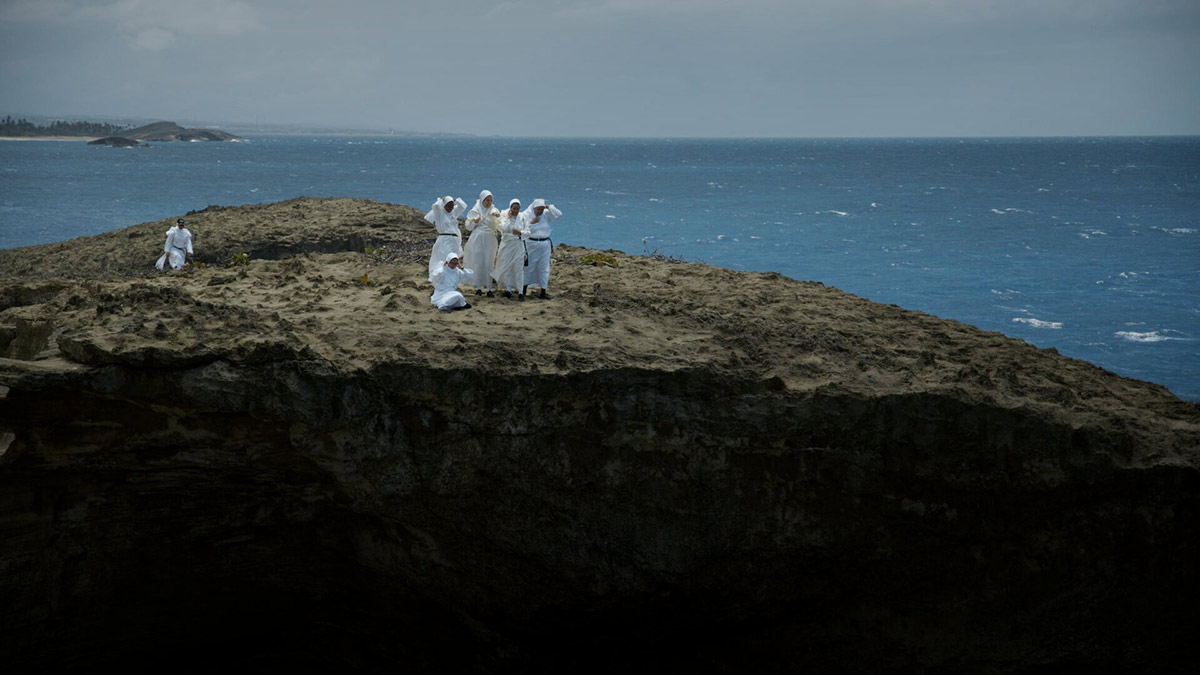 Six nuns dressed all in white stand in the distance on top of a cliff overlooking the ocean