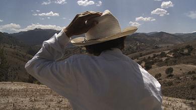 A man with his back to the camera stands and looks out into mountains, holding a fedora on his head