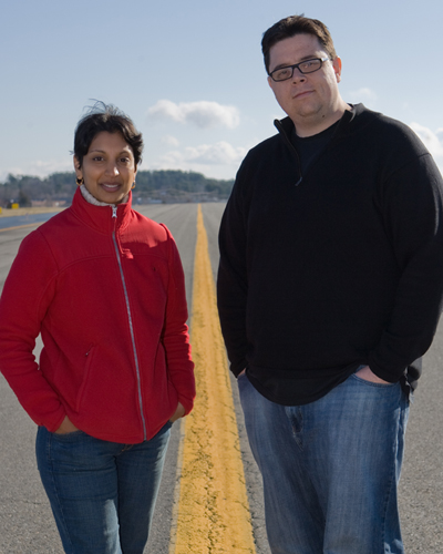 A woman and man standing outside on a road, looking at the camera with their hands in their pockets
