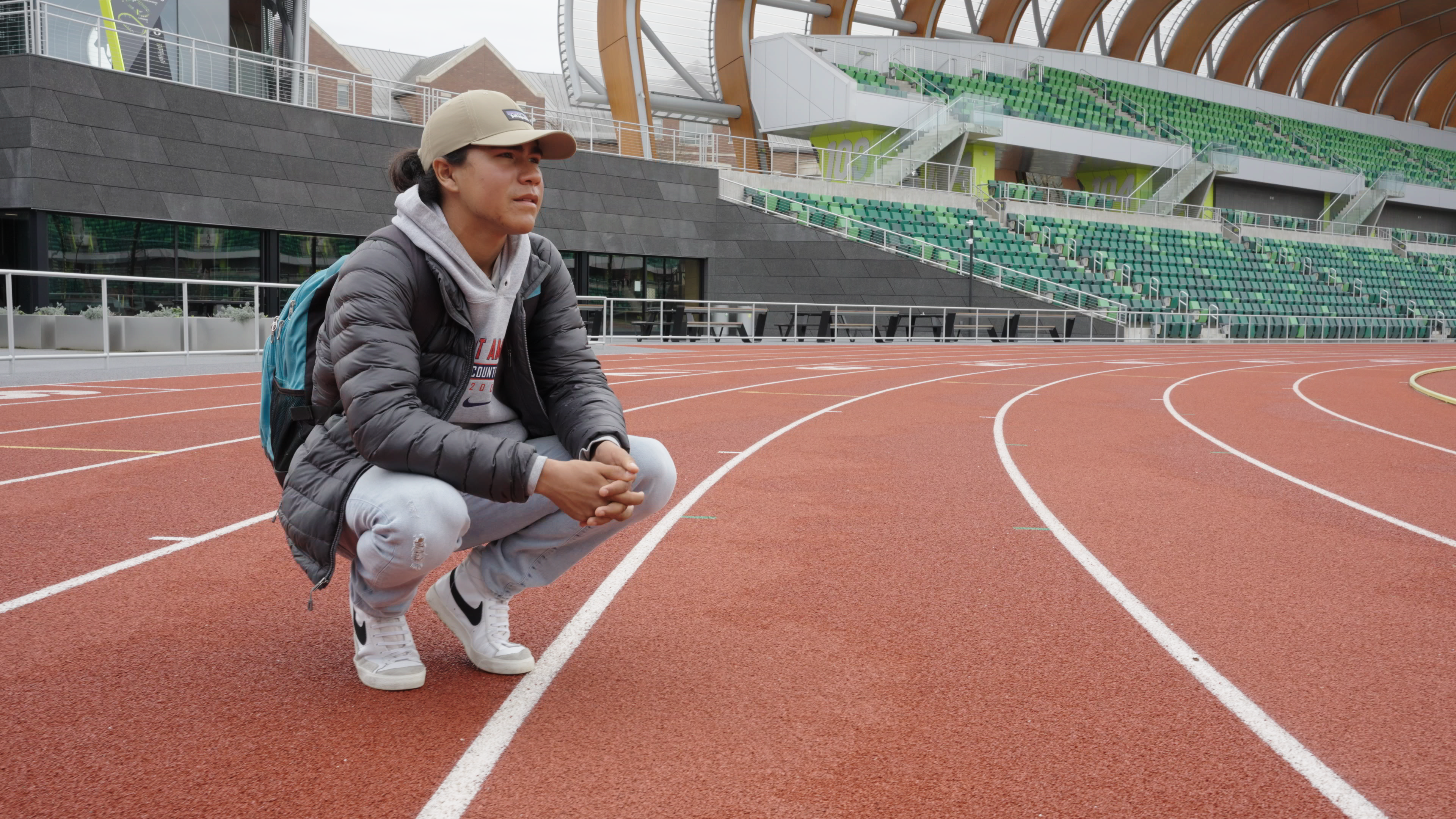Ku, an Indigenous teenager, squats on a running track. He wears a black parka, a beige baseball cap, white pants and running shoes. From Paige Bethmann's ‘Remaining Native’  Photo courtesy of ‘Remaining Native.’ 