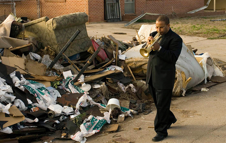 Film still from Spike Lee's 'When the Leeves Broke: A Requiem in Four Acts', featuring a Black man in a suit playing the trumpet while walking next to a trash-filled sidewalk.  