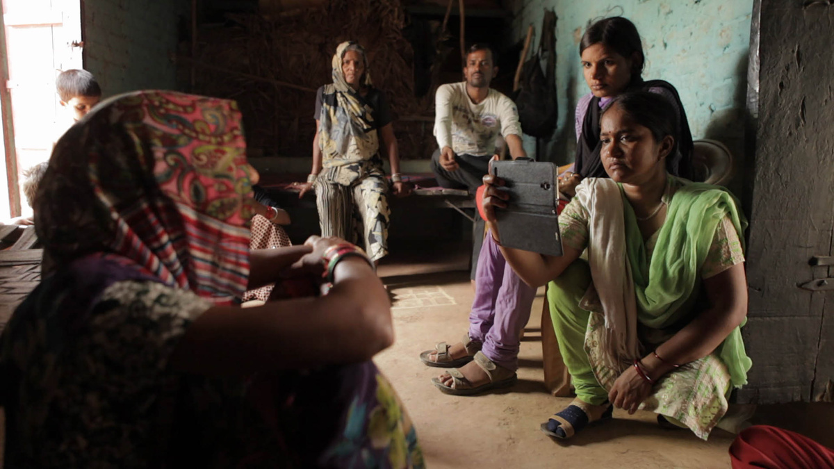 From Rintu Thomas and Sushmit Ghosh's 'Writing with Fire.' Courtesy of Sundance Institute. At right, A reporter from an India-based digital platform is kneeling while interviewing an Indian woman who is seated at left, in her living space with her family. The reporter is filming with a smartphone in her right hand , nd is wearing an emerald green sari