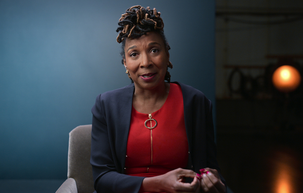 KIMBERLÉ CRENSHAW, Professor, UCLA and Columbia Schools of Law. She is an African-American woman, wearing a red blouse under a navy blue blazer; she is seated against a blue backdrop. From episode 3 of 'AMEND-The Fight for America.' Credit: Courtesy of Netflix/NETFLIX©2021
