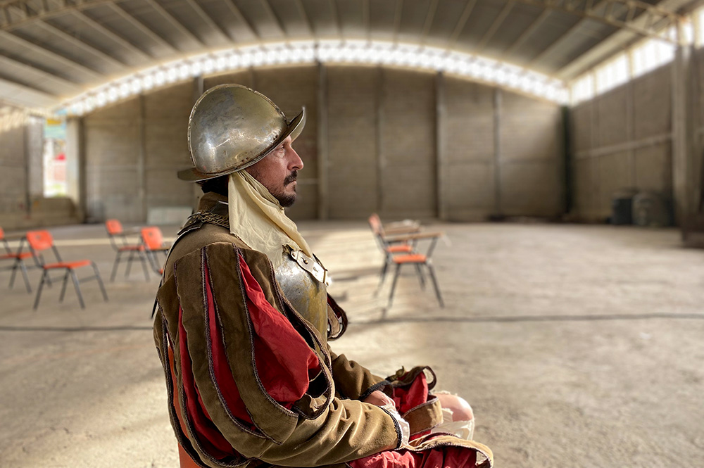 Actor Eduardo San Juan Breña is a bearded Mexican man dressed as a Conquistador. He has a metallic helmet and armor, and is wearing a red and brown costume. Image from Rodrigo Reyes’ “499.” Courtesy of the filmmaker.