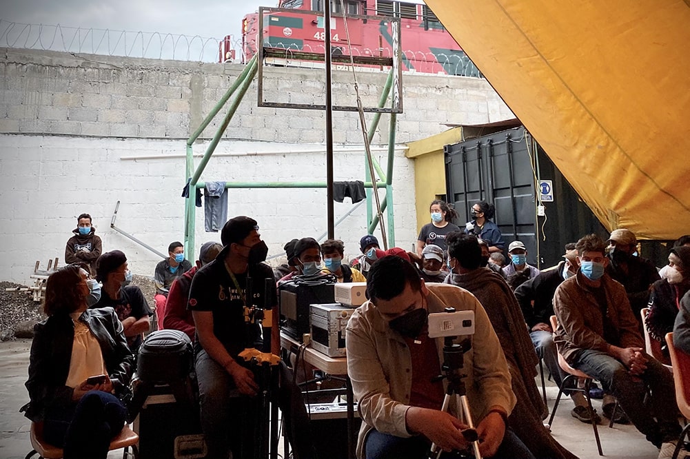 A masked audience gathers under a yellow tarp to watch Rodrigo Reyes’ "499" at the Sacred Family Migrant Shelter. There is a projector setup. Image courtesy of the filmmaker.