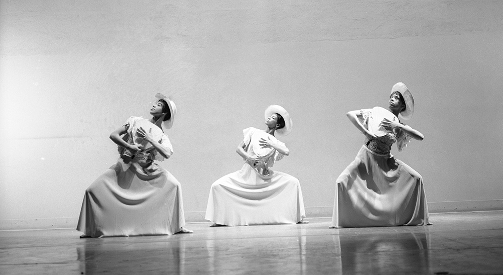Black-and-white image of three Black women dancers holding a pose wearing long dresses, hats, paper fans in their hands. Image from Jamila Wignot’s 'Ailey.' Courtesy of NEON.