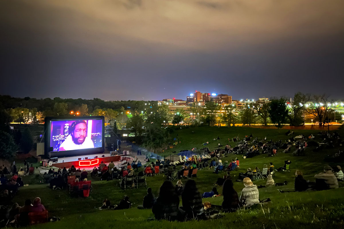 An outdoor screening of ‘Summer of Soul’ at True/False 21. It is night time and people are sitting on chairs and on grass. The projector screen shows the film’s director, Ahmir “Questlove” Thompson, who is a Black man with a beard and short black hair. Courtesy of True/False