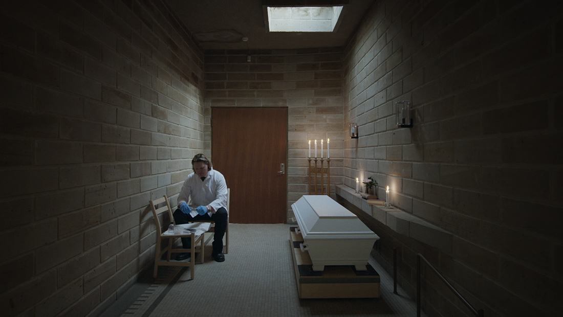 A funerary worker sitting on a chair, next to a closed coffin. There are lit candles surrounding him. A still from 'Meanwhile on Earth,' directed by Carl Olsson. Courtesy of Syndicado Film Sales.