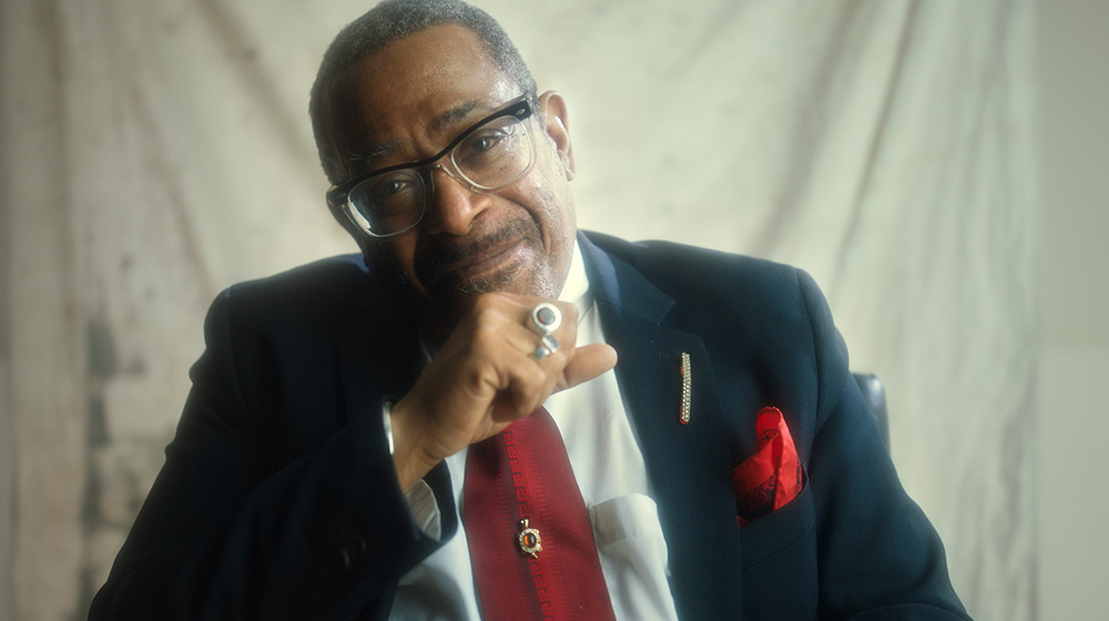 Terence Smith, who performs in drag as Joan Jett Blakk, is a Black man wearing clear glasses, a black suit with a bright red tie and pocket square and two ornate rings. He smiles into the camera. Image from Whitney Skauge's  'The Beauty President.' Courtesy of Outfest.
