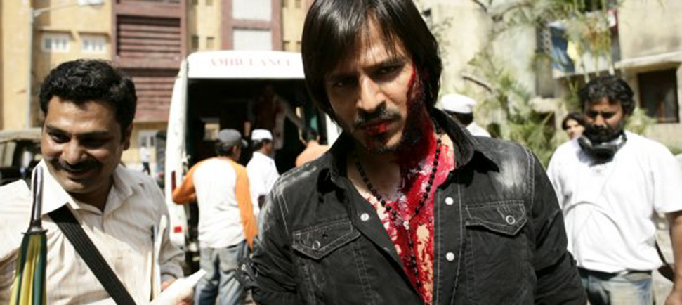 Two men on a movie set. The man in the front has brown skin and is wearing black shirt. He is covered in fake blood. Still from 'Shot in Bombay' by Liz Mermin. Courtesy of IFFLA.