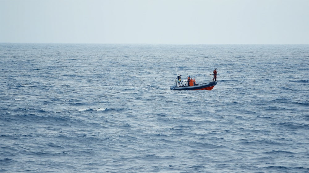 A long-lens shot of the RHIB, an orange-and-grey inflatable boat, amidst a blue sea from the research vessel. Courtesy of Bleecker Street.