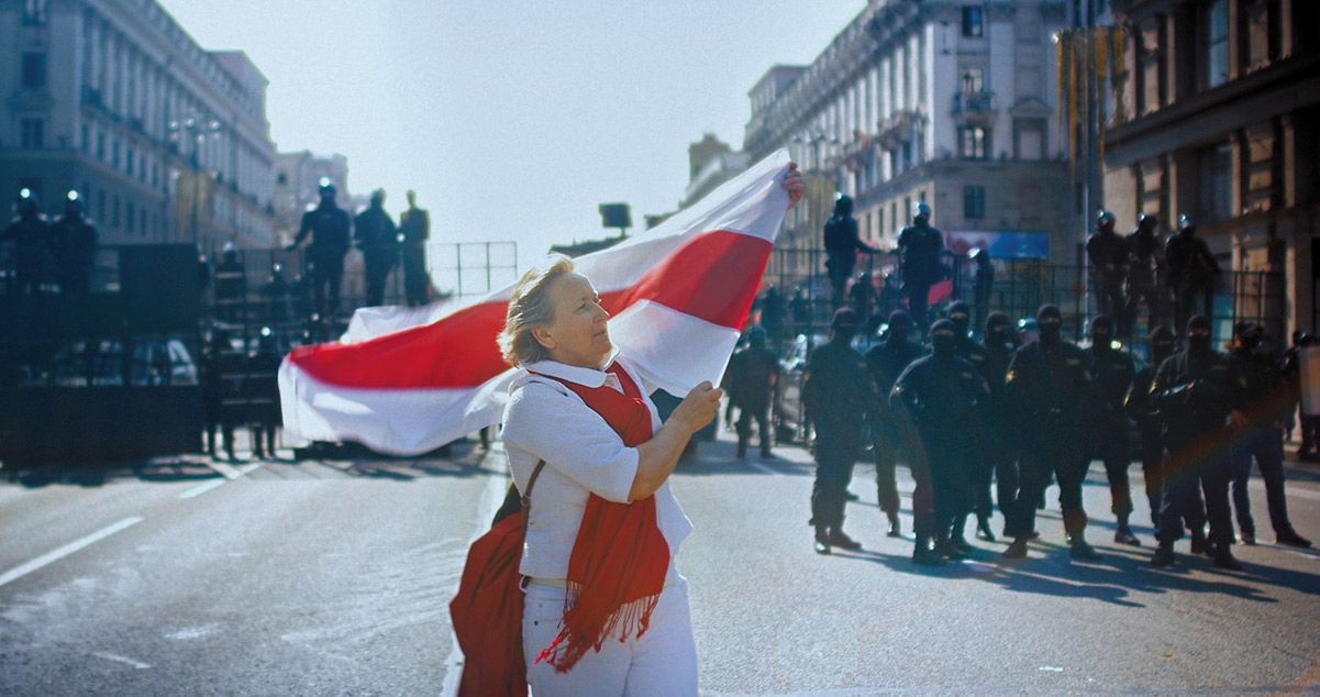 A white woman in a white shirt and pants and a red shawl carries a red and white flag across a city street. From Aliaksei Paluyan’s 'Courage.' Courtesy of CPH:DOX