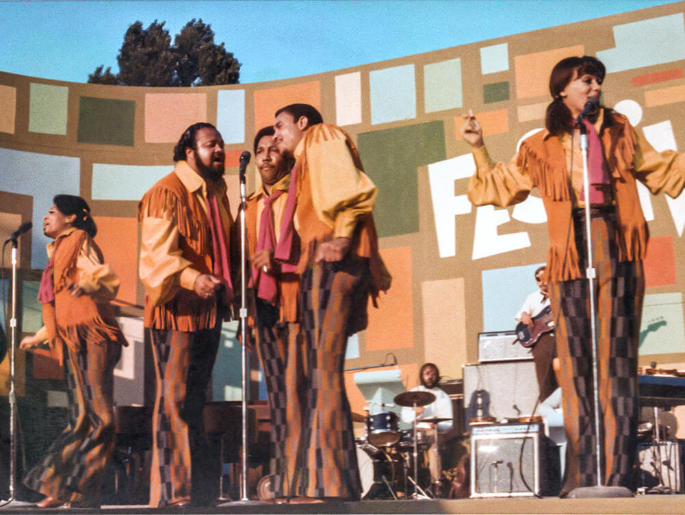 The 5th Dimension, Five singers, two women and three men, in orange and brown outfits singing on a stage, at the Harlem Cultural Festival. Courtesy of Searchlight Pictures.