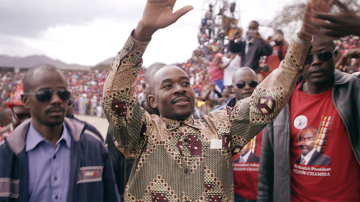 Nelson Chamisa is a bald Zimbabwean male politician waving to a crowd of supporters, surrounded by party workers.  Image from Camilla Nielsson’s ‘President.’ Courtesy of Greenwich Entertainment.