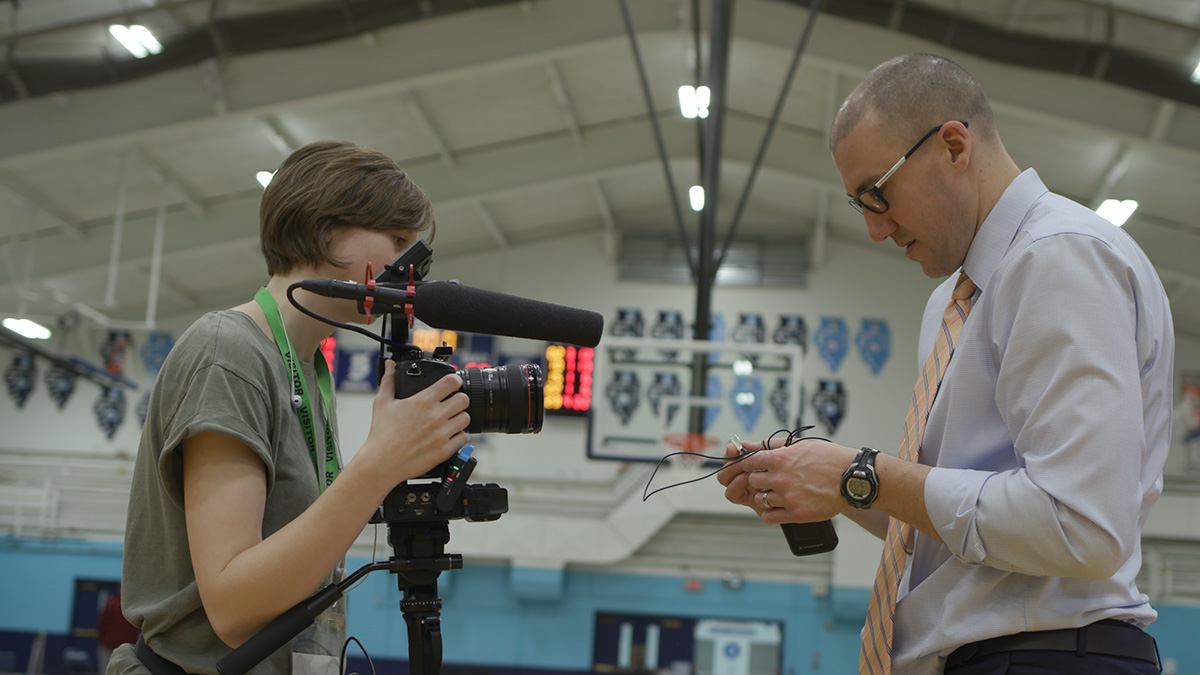 Photo of a woman in her late 20s standing in a basketball gym, with her camera and a male teacher wearing a tie, handing her his microphone.