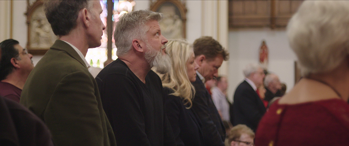 Two white men--Michael Sandridge (in a moss green jacket) and Dan Laurine (in a black shirt)--stand in a church service. From Robert Greene’s “Procession.” Courtesy of Netflix.