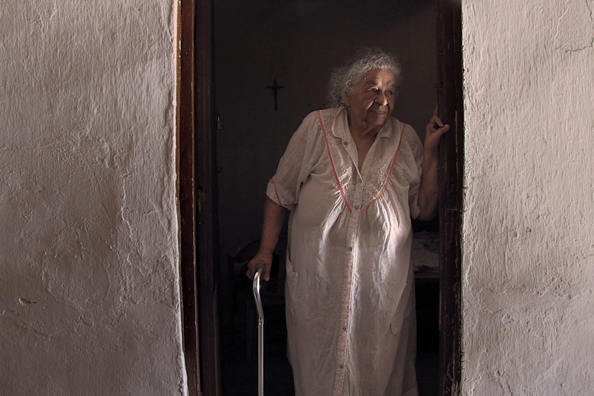  Mama Icha is a senior Colombian woman seen here standing in her nightgown, holding a cane. From Óscar Molina’s ‘La Casa de Mama Icha.’ Courtesy of Óscar Molina.