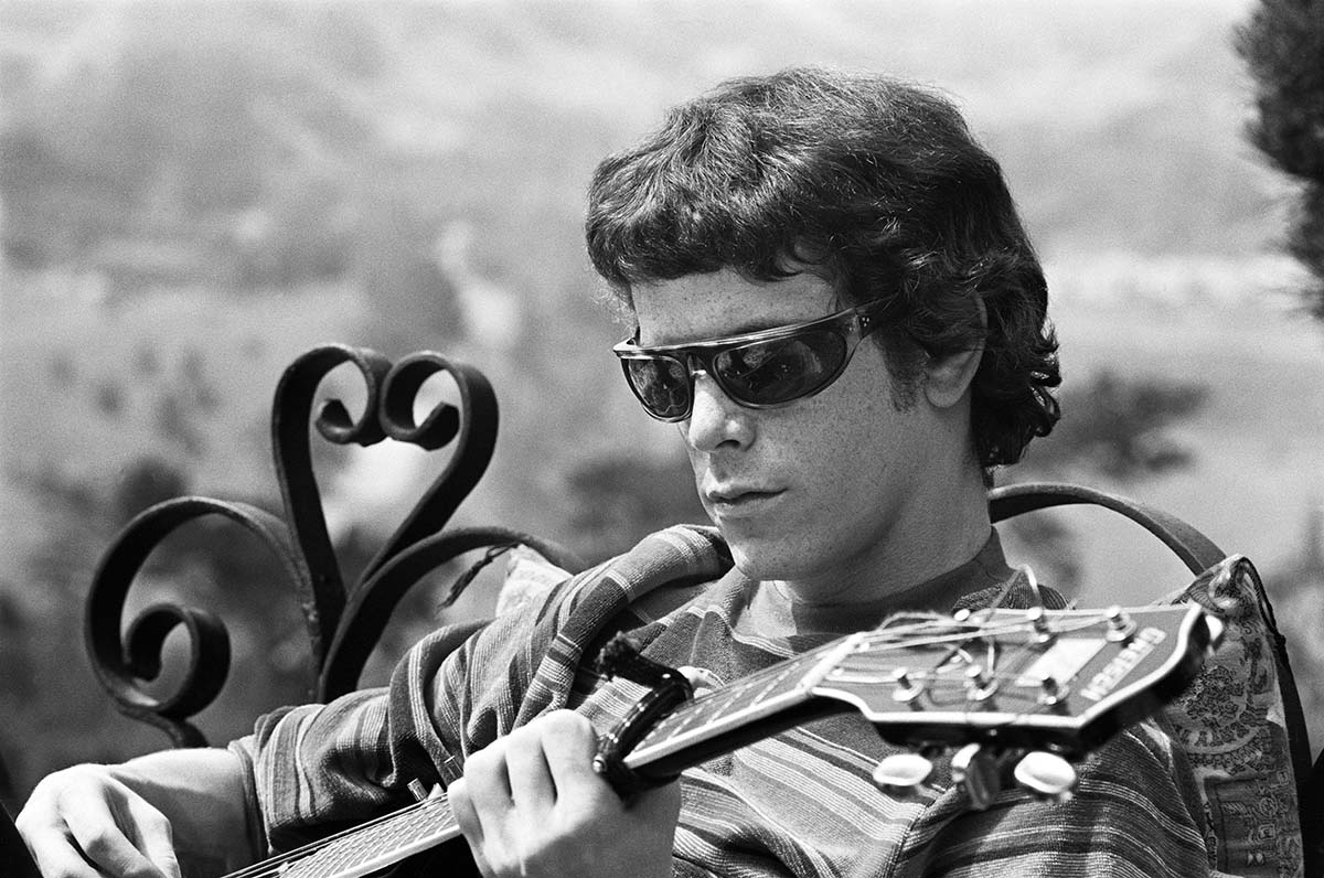 Lou Reed, a white male with black wraparound shades and black hair and a striped shirt, sits on a bench playing a guitar. From Todd Haynes' 'The Velvet Underground.' Courtesy of Apple TV.