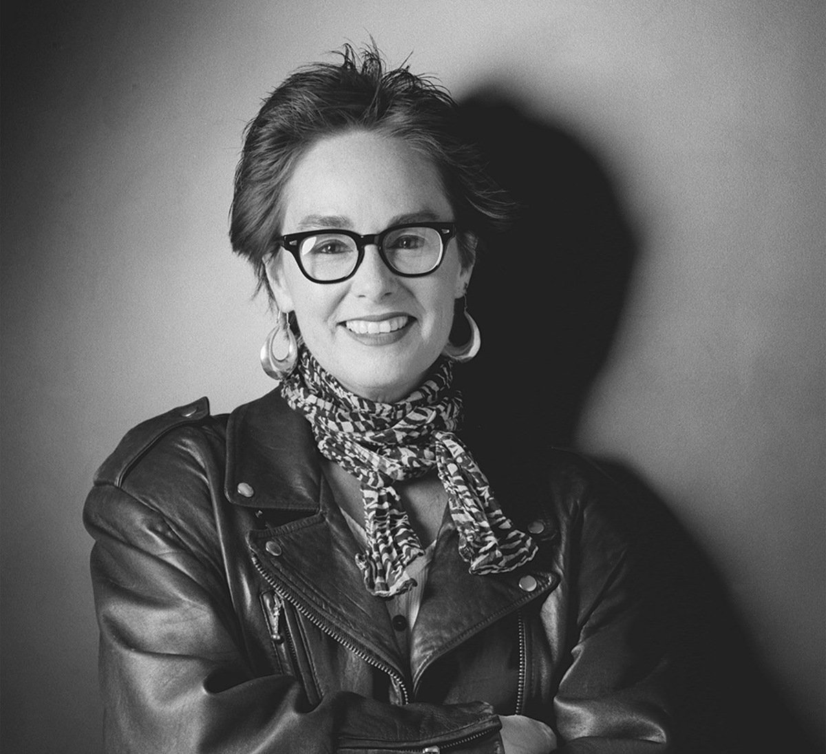 The late filmmaker/educator Judy Irola. Photo: Douglas Kirkland. Courtesy of American Society of Cinematographers. In this black-and-white portrait of Judy Irola, she has short brunette hair and glasses, she is wearing a leather jacket and she is posing with her arms crossed across her chest.