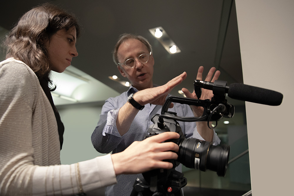 Bob Sacha, a white male professor, working with a young female student at Newmark J School. Both of them are looking at a camera fitted with a microphone. Photo by Marco Poggio.