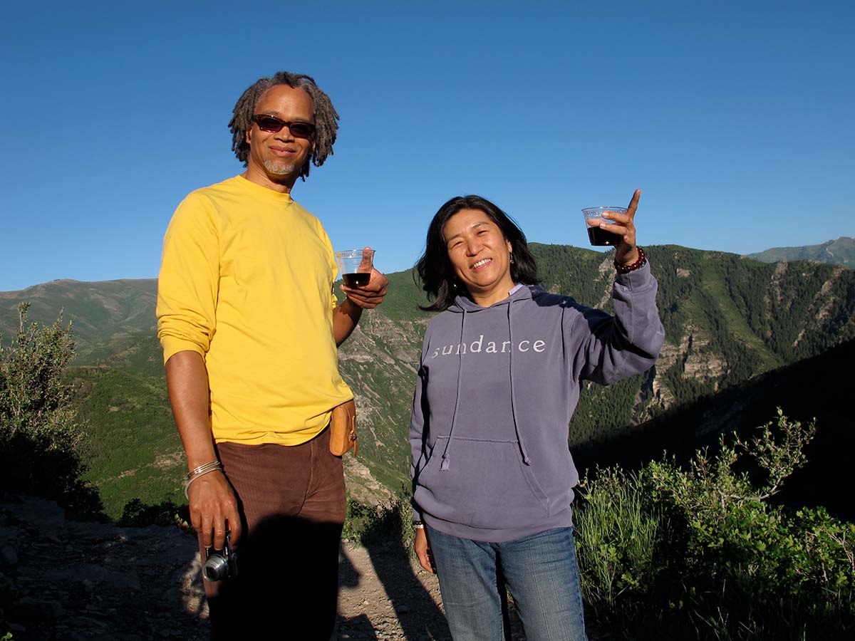Asian American filmmaker Jean Tsien raising a toast with legendary Black male editor Lewis Erskine, with green mountains surrounding them. Photo courtesy of Jean Tsien.