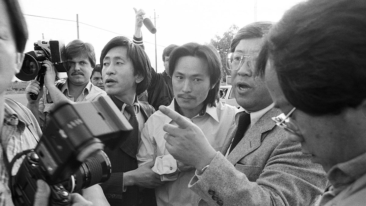 Choi Soo Lee is an Asian man in a white short; he is surrounded by journalist and camerapeople. From 'Free Chol Soo Lee' by Julie Ha and Eugene Yi, photo by Grant Din.