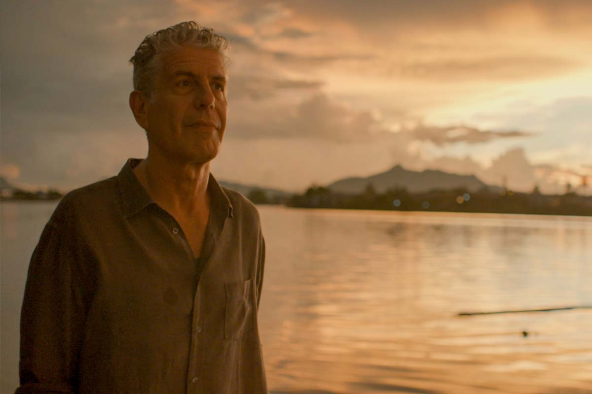 Anthony Bourdain, a white man with white hair,, wearing a button-down shirt, stands on a beach, with the sun setting behind him.  From Morgan Neville's 'Roadrunner.' Courtesy of CNN / Focus Features. 