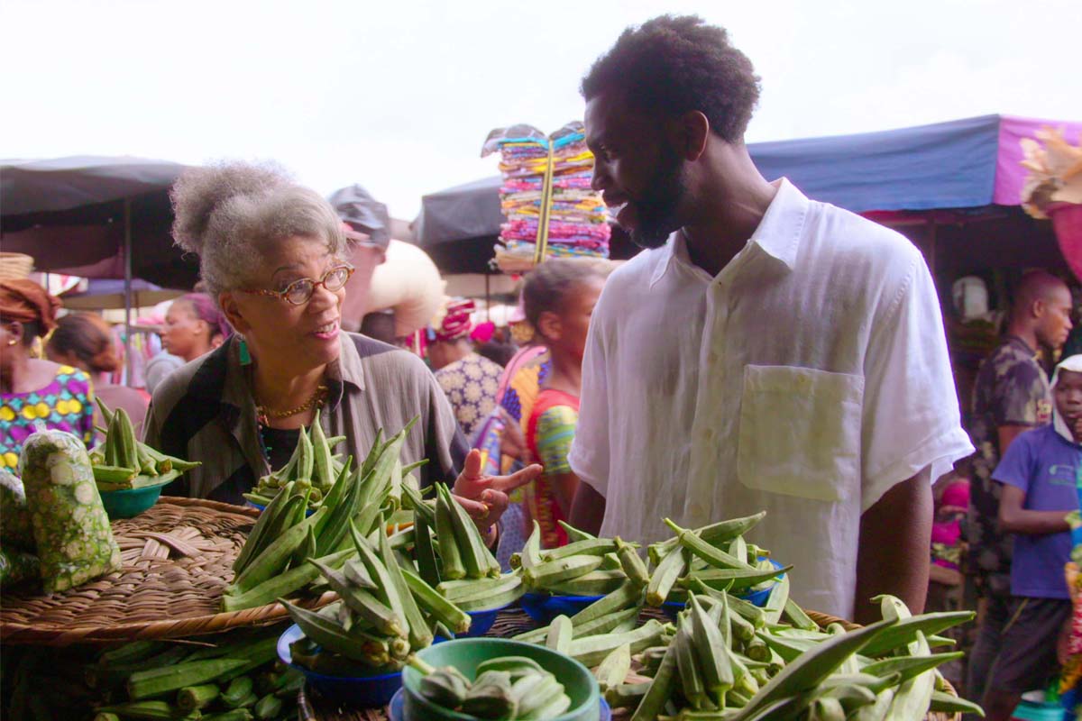 Dr. Jessica B. Harris, a Black female historian, stands with Black male writer Stephen Satterfield in a fresh produce marketplace in Benin, Africa. From 'High on the Hog' co-directed by Roger Ross Williams. Courtesy of Netflix. 