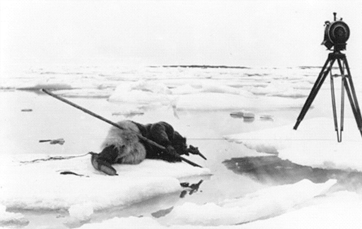 A camera sits on an ice sheet while a man hunts while laying down with a spear facing towards the water nearby. From Robert J. Flaherty's 'Nanook of the North.'