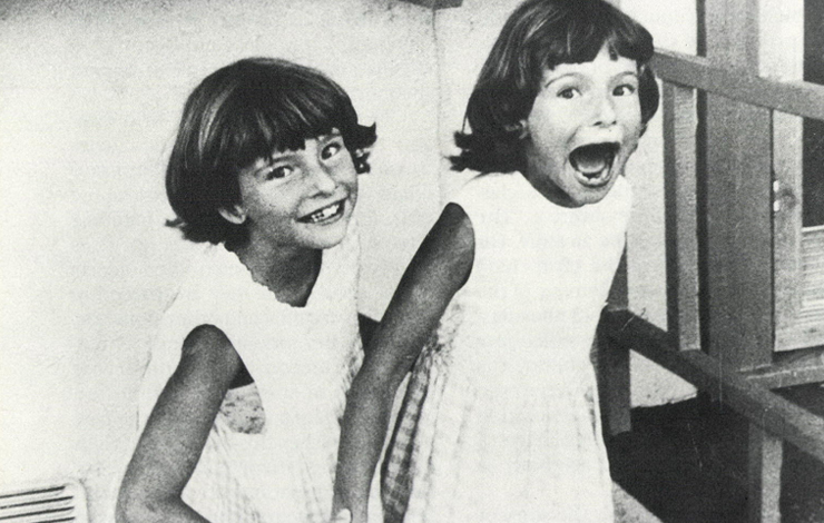 Two twin girls with bangs and short hair; one smiles, while the other ones mouth is agape as if shouting. From 'Poto and Cabengo.' 