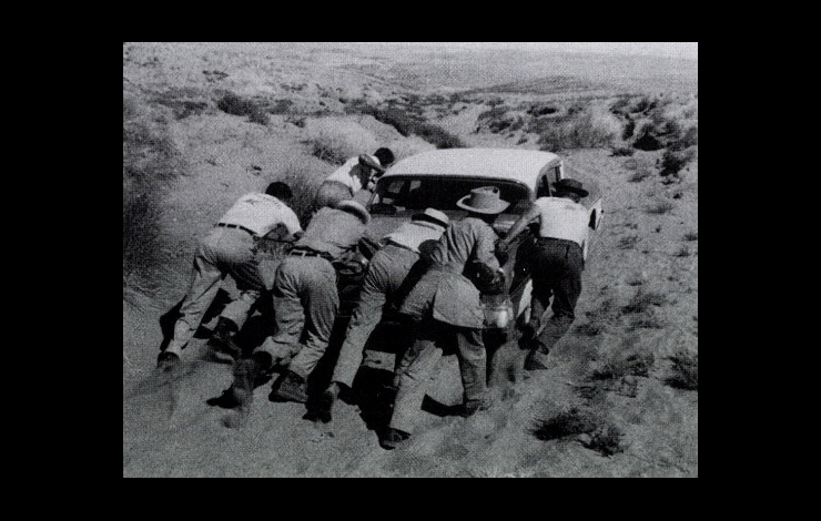 A black-and-white photo of a group of people pushing a car through a barren landscape.. 