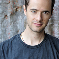 Headshot of an adult male with light skin tone, short brown hair, in a black tee shirt, in front of a weathered, corrugated metal wall.