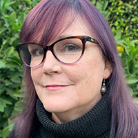 Headshot of a mature woman with pale skin and purple shoulder length hair. She has brown eyes and wears large purple glasses and a black turtleneck sweater.