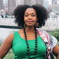Headshot of a stunning black queen, her natural curly hair billowing like a crown in an emerald green tank top with the great city of New York as her backdrop.
