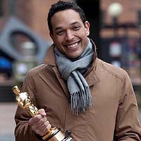 A mixed-race man wearing a brown coat and grey scarf holding an Oscar award.