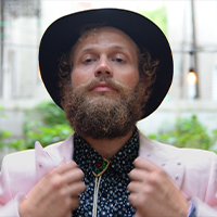 Bearded white beauty in a pink blazer and a black cowboy hat, adjusts their lapel while looking dashingly into the camera. 