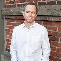 A white man in a white shirt in front of a brick building.