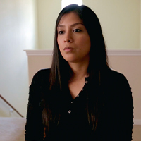 A Latina with dark, straight hair sitting in a room with white walls and a staircase in the background.