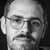 A black and white picture of a man with light skin and dark short hair and facial hair with round glasses.