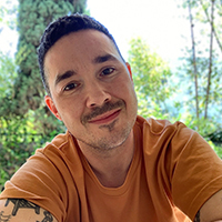 A trans man with brown hair, smiling and wearing an orange shirt.