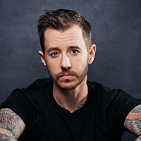 A trans man with brown hair, wearing a black shirt, sitting with his arms clasped.