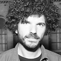 A black and white portrait of a wan with light skin and curly black short hair and facial hair.