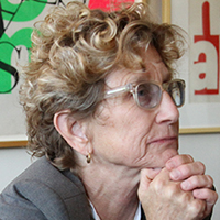 An older woman with light skin tone, blonde curly hair and round glasses.