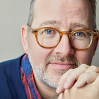 A man with brown, round glasses and gray hair and facial hair.