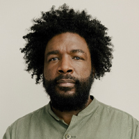 A Black man with black curly hair and facial hair.
