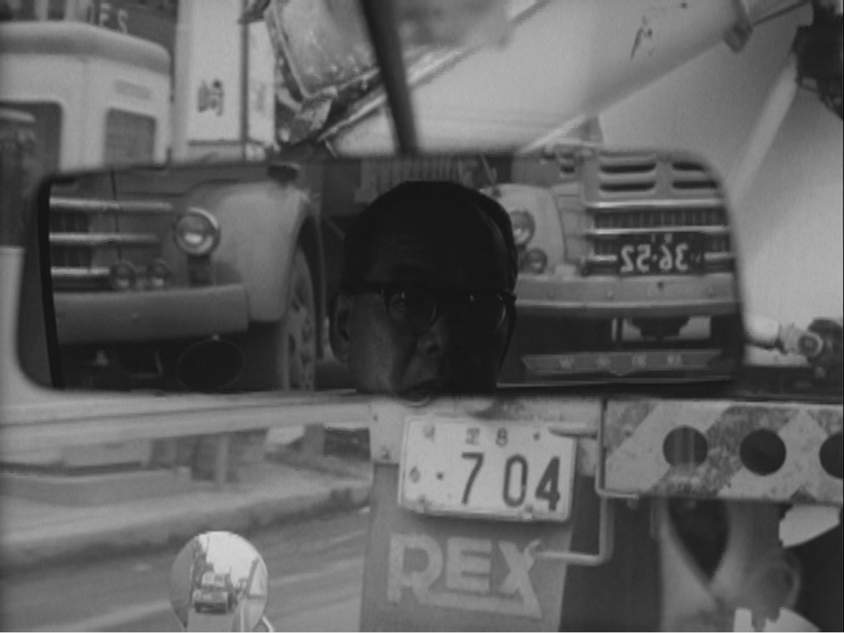 A man’s reflection in a rearview mirror, from Noriaki Tsuchimoto’, ‘On the Road: A Document’ (1964). Photo courtesy of Motoko Tsuchimoto. 