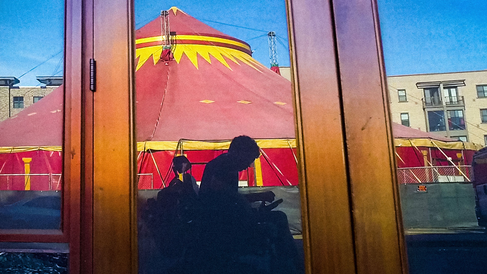 In a reflection of an unmarked storefront is a grayish silhouette of a man using an electric wheelchair. Behind the man is a spectacular red and yellow circus tent. Photo courtesy of Reid Davenport.