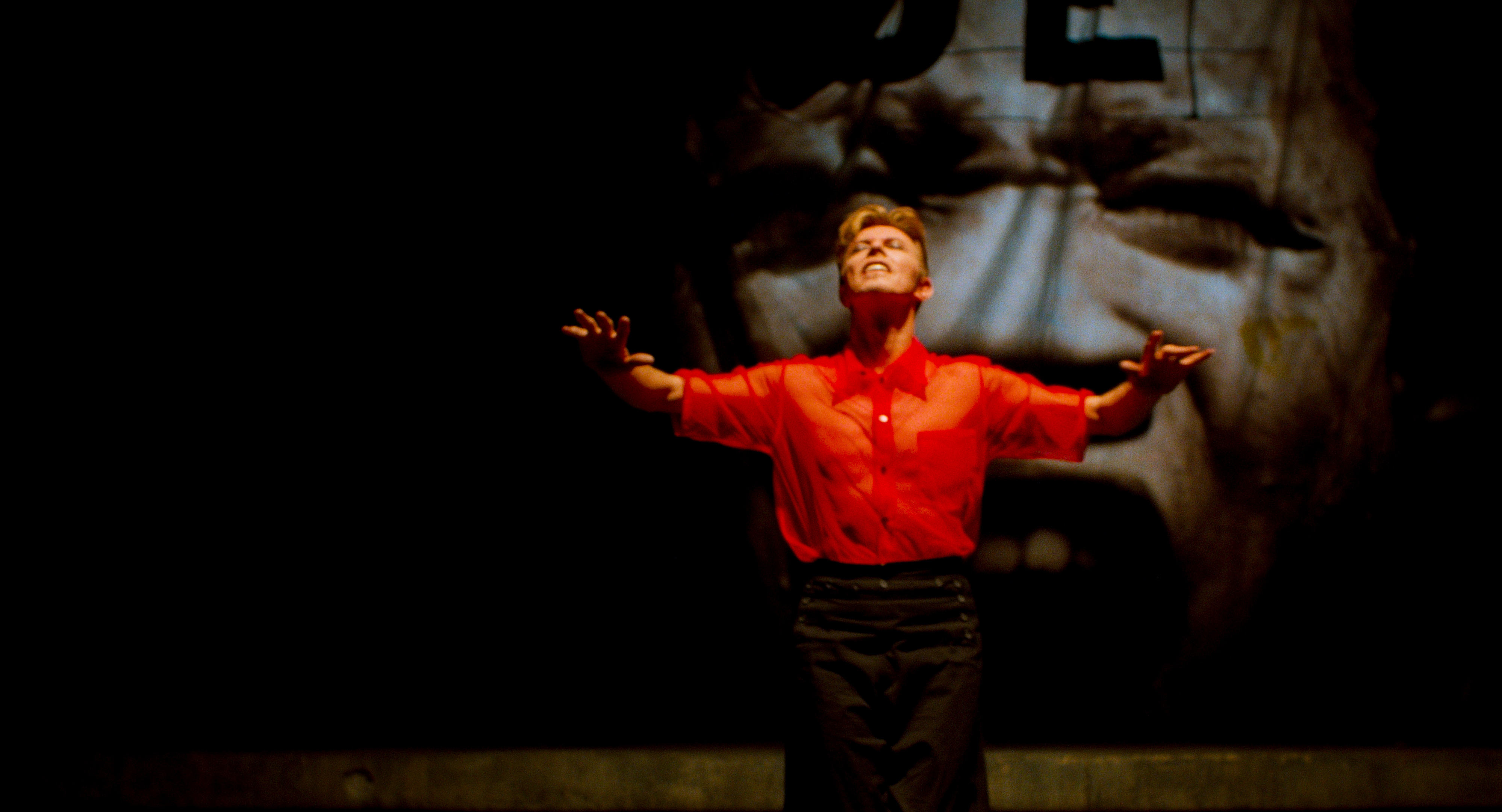  David Bowie stands with his eyes closed and arms up in front of a projection of someone’s face, from ‘Moonage Daydream.’ Photo courtesy of NEON. 