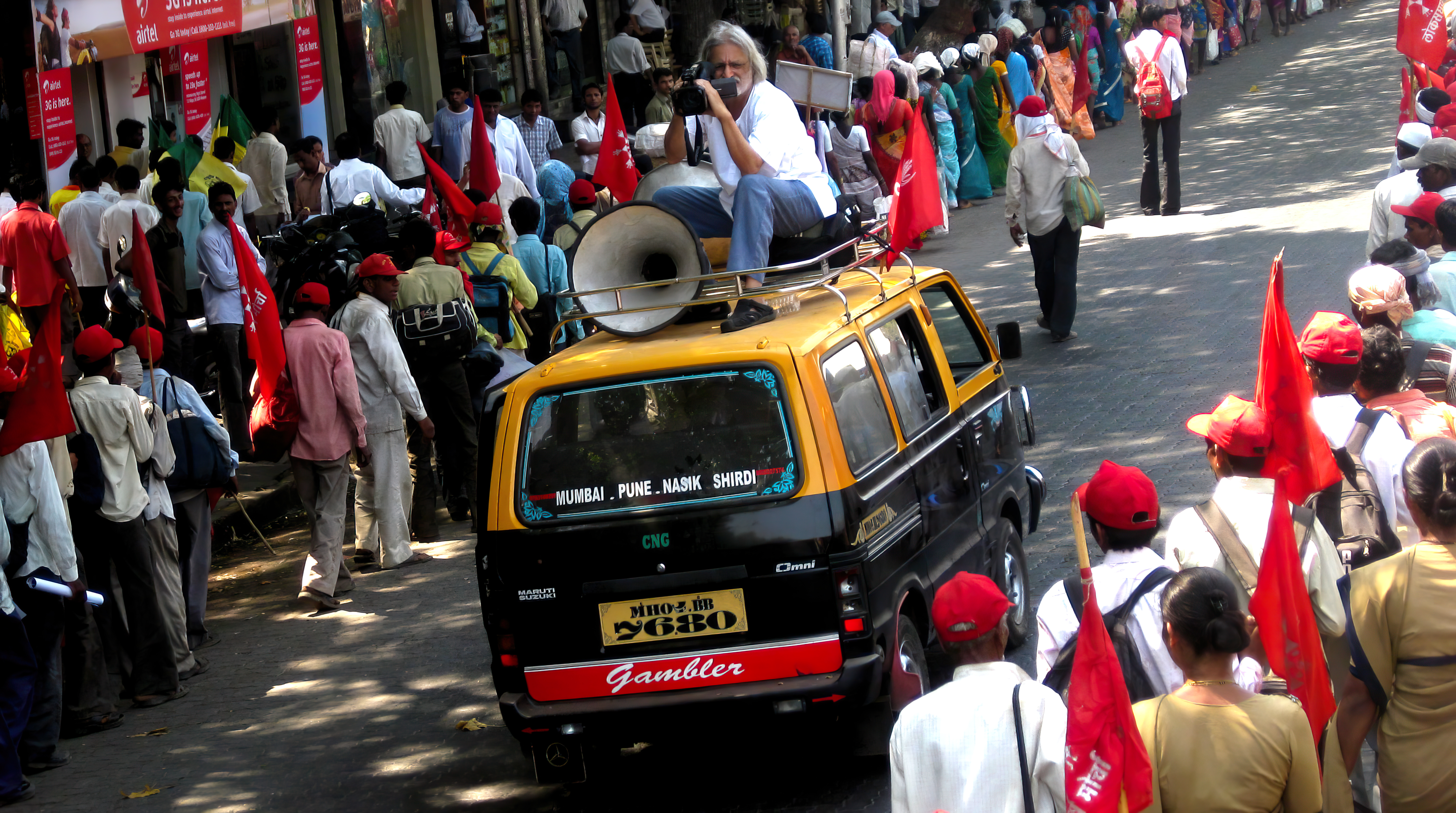 Anand Patwardhan, an Indian man with long white hair, seated atop a van that is moving through the street, is filming 'Jai Bhim Comrade' (2011). Courtesy of Anand Patwardhan. Photo: Priyanka Borpujari .