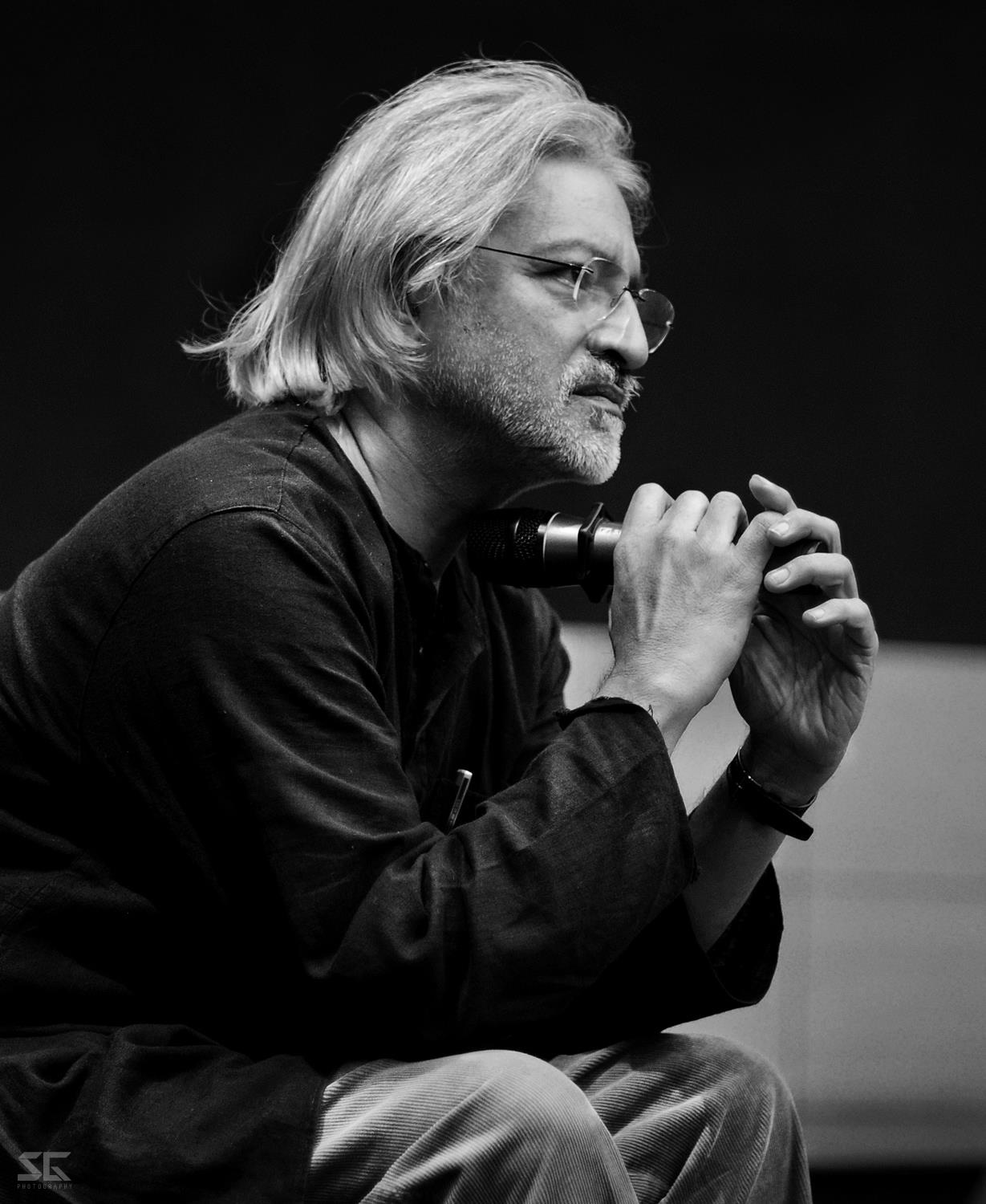 A black-and-white headshot of Anand Patwardhan, an Indian man with long, white hair and glasses and a black shirt; he holds a microphone., wearing a black shirt.. Courtesy of Anand Patwardhan. Photo: Sagnik